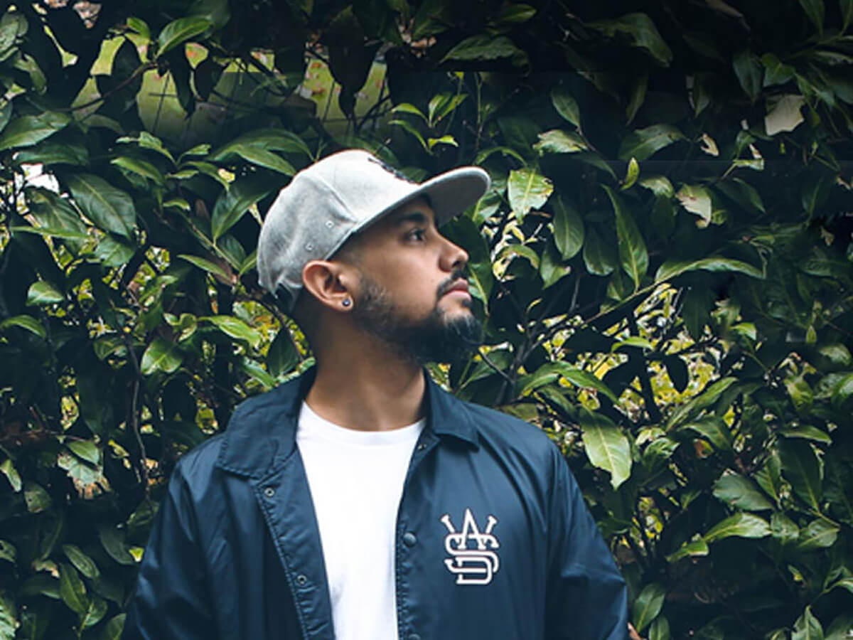 A male model wearing a blue WASD jacket in front of a green hedge.