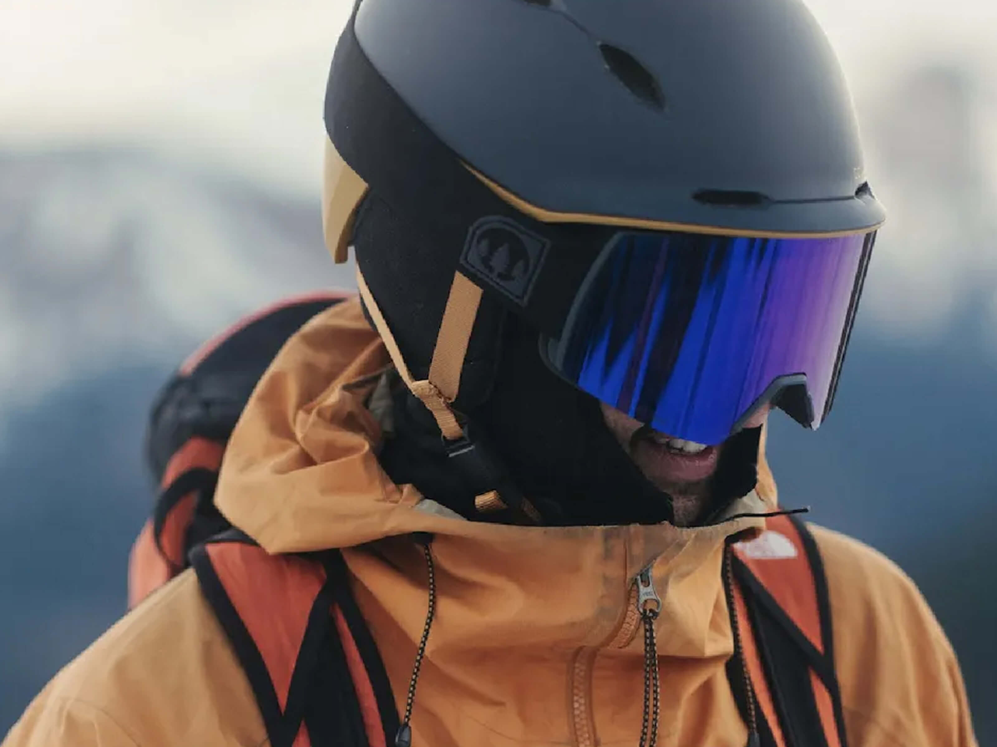 Skier wearing a black helmet and blue mirrored ski goggles with an orange jacket on a mountain.