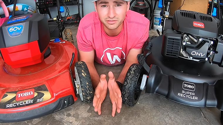 A man laying on his stomach on a garage floor between two mowers, one red and one black.