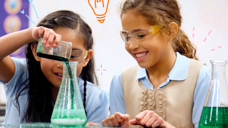 Two girls wearing safety glasses, one pouring liquid into a beaker and the other smiling and watching.
