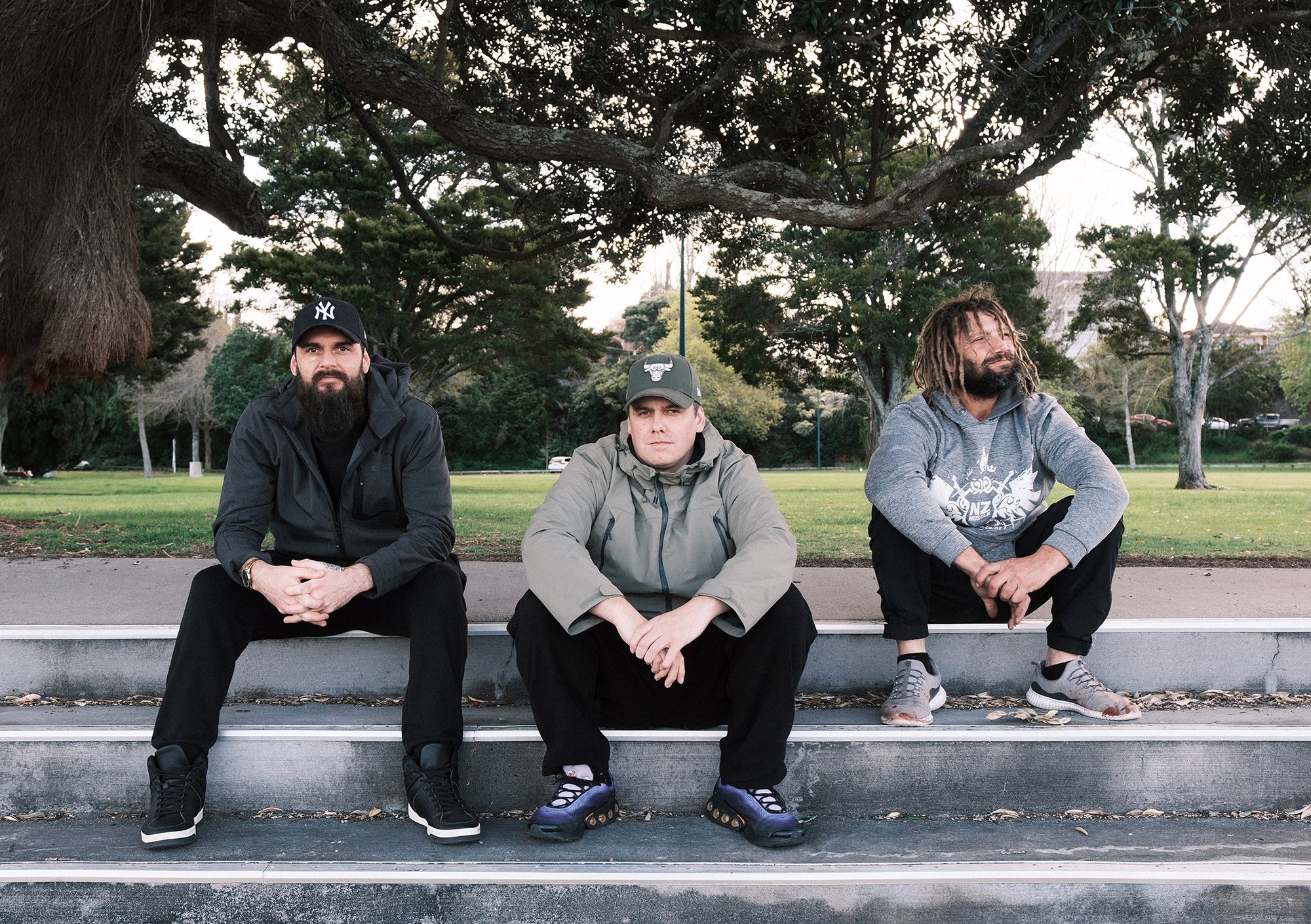 Adam Lilburn, Carl Fenton and Aaron sit on concrete steps beneath a large tree in a park