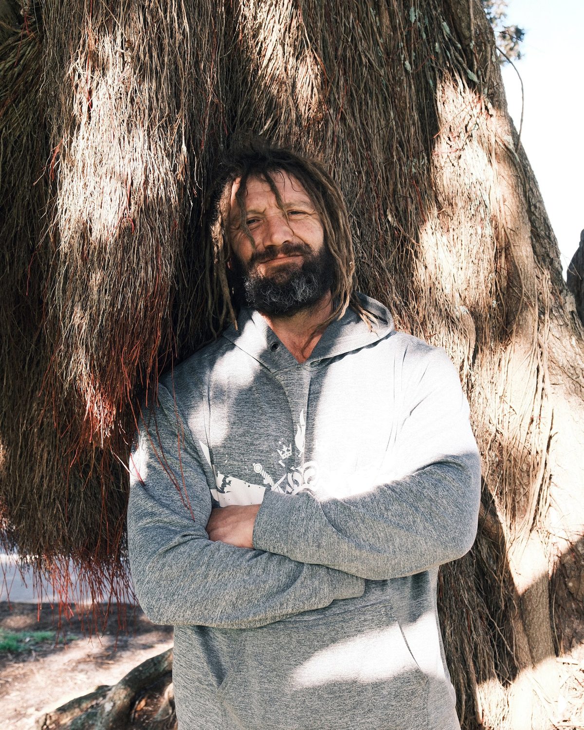 Aaron in a grey hoodie stands with his arms crossed in front of a large, shaggy tree trunk