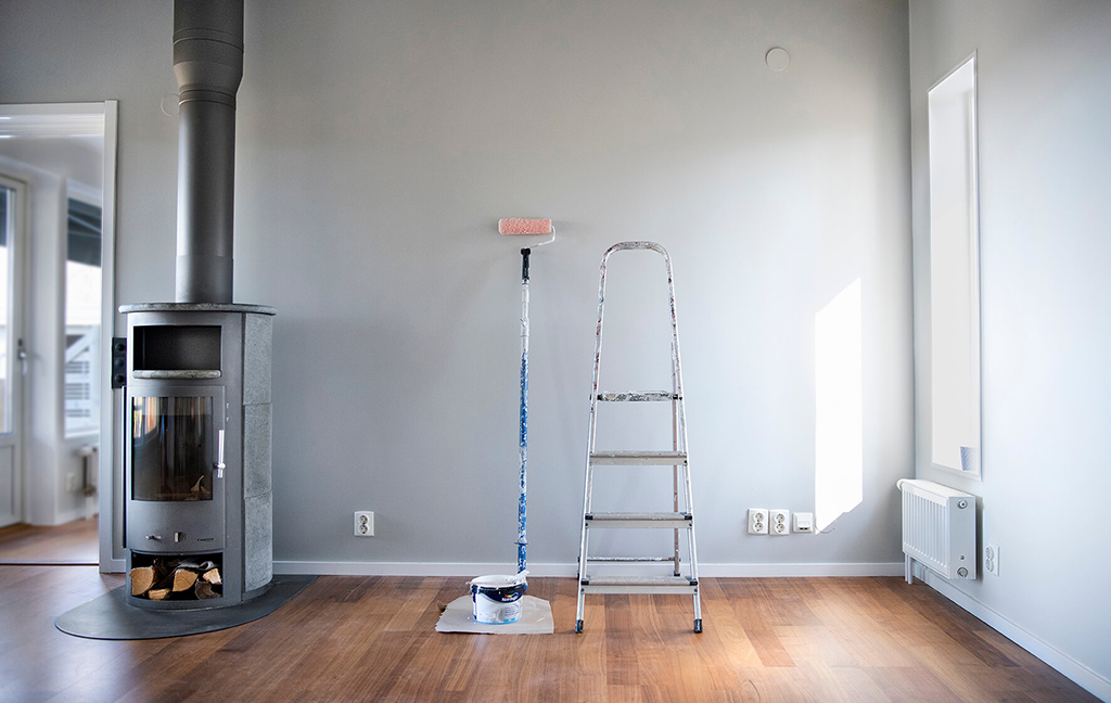 Eldstad med ved, målarstege och målarroller mot en grå vägg i ett rum med trägolv.