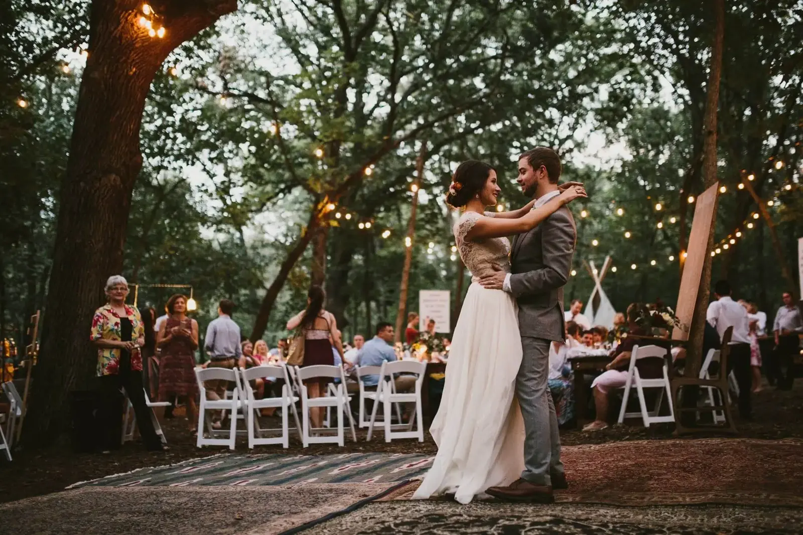 Bride and groom embracing and dancing outdoors at a wooded wedding reception with string lights and seated guests in the background.