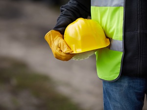 A construction worker wearing gloves and a safety vets, holding a hardhat.