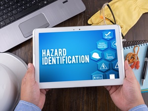 Person holding a tablet displaying the words "Hazard Identification" with various safety icons on the screen, surrounded by safety equipment such as gloves, hard hat, and notebook on a desk.