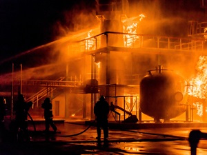 Firefighters responding to a large industrial fire at a facility. Flames engulf structures and tanks, highlighting a hazardous chemical incident requiring emergency response.