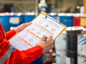 A worker in a red uniform holding a clipboard with a checklist for chemical safety in an industrial setting. Various containers and safety labels are visible in the background.