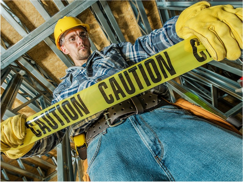 A construction worker in safety gear holding yellow caution tape inside a building under construction.