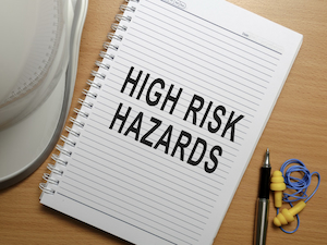 A spiral notebook on a desk with the words “High Risk Hazards” written on the page, next to safety gear including a hard hat, earplugs, and a pen.