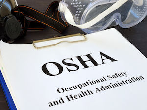 A clipboard with the word “OSHA” and the text “Occupational Safety and Health Administration,” placed next to protective goggles and a respirator strap.