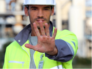 Construction worker in a high-visibility jacket and hard hat holding up a hand in a stop gesture, signaling to pause or stay back.
