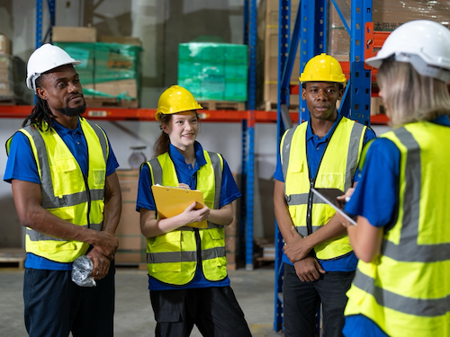 A diverse group of warehouse workers wearing high-visibility vests and hard hats listen attentively to a supervisor during a safety briefing.