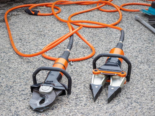Two hydraulic rescue tools with orange hoses lie on the pavement, used for cutting and spreading during emergency or industrial operations.