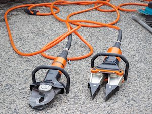 Two hydraulic rescue tools with orange hoses lie on the pavement, used for cutting and spreading during emergency or industrial operations.