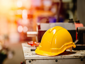 A yellow hard hat rests on top of industrial equipment in a workshop, with blurred tools and machinery in the background.