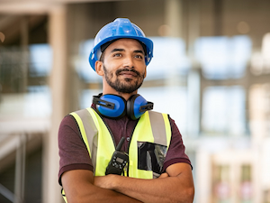 A male worker wearing a blue hard hat, hearing protection, safety vest, and a two-way radio stands confidently in a bright indoor work environment.