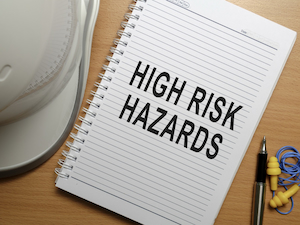 A lined notebook on a desk displays the words “HIGH RISK HAZARDS” next to a white hard hat, earplugs, and a pen.