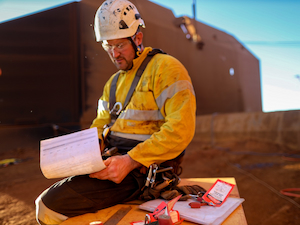 Construction worker wearing yellow protective gear and helmet, sitting outdoors while reviewing safety paperwork and equipment tags.