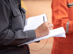Worker in dark clothing writing notes in a safety logbook while another person in orange stands nearby.