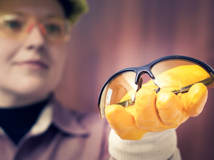Worker wearing gloves holding up a pair of amber-tinted safety glasses for inspection.