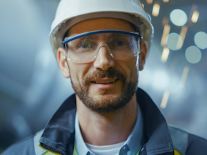 Smiling worker in a hard hat and clear safety glasses standing in an industrial environment.