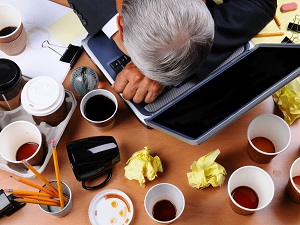 A business person with his head on his computer and multiple coffee cups and trash all around him because he is stressed and tired but his desk is very messy.