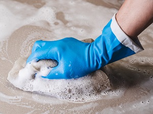 A hand wearing a rubber glove cleaning a hard surface with soapy water.