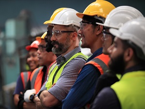 Several manufacturing plant employees standing in a row all wearing safety glasses, hardhats, and safety vests.