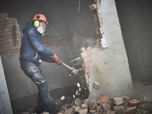 A worker wearing PPE using a sledgehammer to tear down a brick wall.