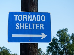 A blue sign on a pole outdoors that says Tornado Shelter with an arrow pointing to the right.
