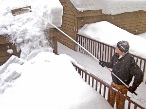 A worker using a snow rake from a lower level to remove snow from a roof.