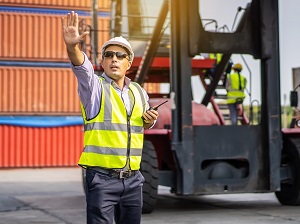 Worker on a job site wearing a safety vest and holding a walkie-talkie with his hand in the air indicating for an action to stop.
