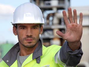 A worker on a job site earing a safety jacket and a hard hat with his hand held out in the universal gesture for stop.