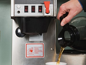 Worker pouring coffee into a cup from a large coffee maker in an office break room.