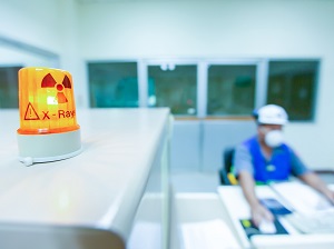 A worker sitting at a desk, wearing a face mask, near an x-ray machine.