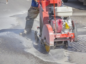 Construction worker wearing rubber boots operating a walk-behind saw to cut into the road.