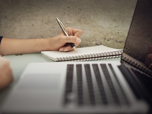 Woman using a pen and notepad to take notes next to an open laptop computer.