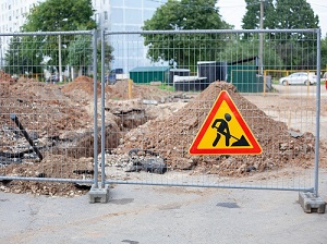 Outdoor construction site where trenching is occurring surrounded by a chain link fence and a hazard warning sign.