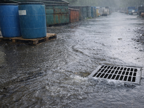 Rainwater runoff flowing across an industrial yard toward a storm drain near storage barrels.