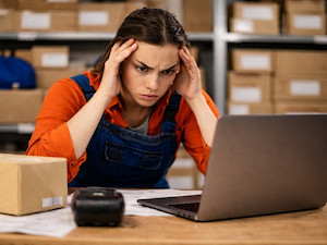 Stressed warehouse employee holding her head while looking at a laptop surrounded by boxes and paperwork.