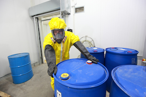 Worker in chemical protective suit and gloves handling hazardous material drums.