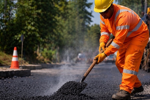 Construction worker in high-visibility clothing shoveling hot asphalt during road paving operations.