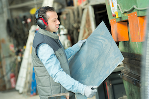 Worker operating a press machine handling sheet metal while wearing protective gloves and hearing protection.
