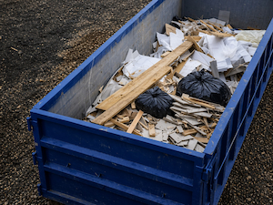 Blue dumpster filled with construction debris including wood scraps, drywall, and trash bags at a jobsite.