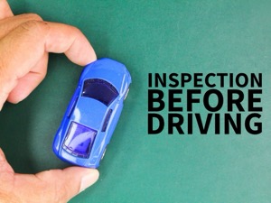 A close-up of a hand holding a small blue toy car against a green background. The text overlay reads: “INSPECTION BEFORE DRIVING.”
