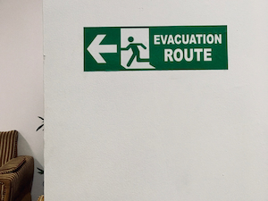 Green “Evacuation Route” sign on a white wall with a left-facing arrow and pictogram of a person exiting through a door.