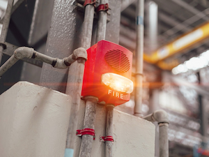 Red fire alarm with a flashing light mounted on an industrial wall with metal conduit pipes in the background.