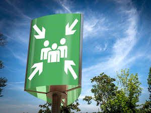 Green sign outdoors with arrows pointing toward a group of people icon, indicating a designated assembly or muster point.