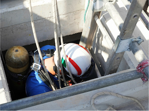 Worker wearing a harness entering a confined space using a ladder.